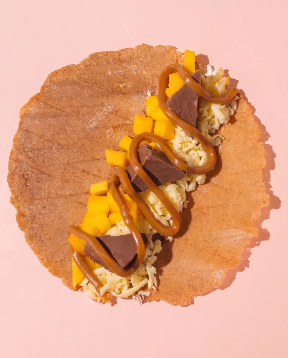 Oblea de marquesita extendida con queso al centro, mango al lado y manjar en zigzag encima, antes de enrollar
